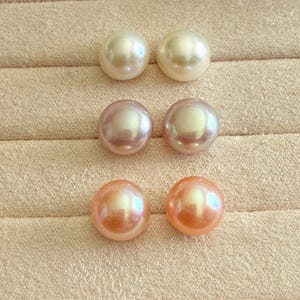 Pode incluir: Seis pérolas redondas e brilhantes em tons de creme, rosa e pêssego. As pérolas estão dispostas em pares, mostrando suas superfícies lisas e iridescentes. As pérolas são exibidas em uma superfície bege.
