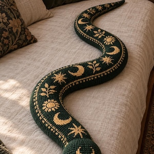 May include: A long, stuffed snake-shaped cushion in dark forest green with a crocheted texture. The body features repeating yellow embroidered patterns of crescent moons, suns, stars, and leafy sprigs. It has two small, round black eyes.