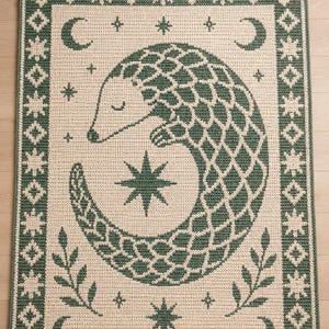 May include: A rectangular crochet rug featuring a cream-colored background with a dark green border and a central graphic of a curled pangolin. The design includes celestial motifs like crescent moons and stars, along with small leaf sprigs.