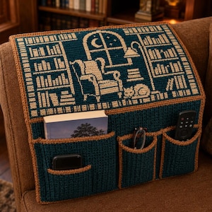 May include: A dark teal and tan crochet armchair caddy featuring a tapestry design of a library with a chair, window, and cat. The organizer includes three front pockets containing a book, eyeglasses, a remote control, and a smartphone.