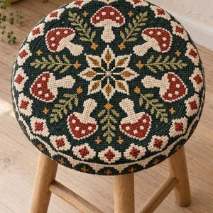 May include: A round wooden stool featuring a needlepoint seat cover with a dark green background. The design includes a central star-like floral motif surrounded by repeating mushroom and fern patterns in shades of red, cream, and gold.