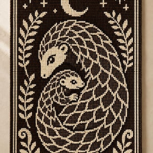 May include: A rectangular wall hanging featuring a dark brown and cream crochet design of a parent and baby pangolin. The scene includes a crescent moon, stars, and leafy branches framing the animals, all suspended from a wooden dowel rod.