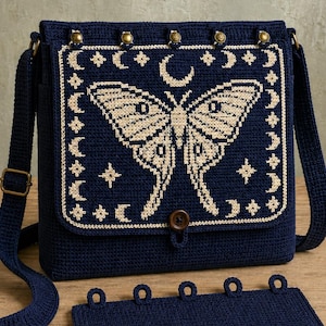 May include: A dark blue crochet crossbody bag featuring a cream-colored moth and celestial pattern on the front flap. The bag has a single brown button closure, an adjustable shoulder strap, and five brass-toned metal studs along the top edge.