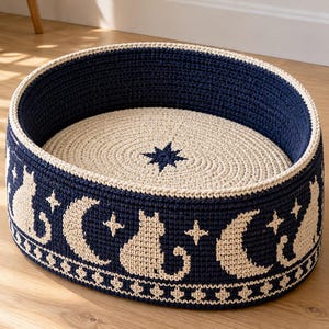 Op de afbeelding: Een rond, gehaakt huisdierbed met een donkerblauwe buitenkant en een herhalend crèmekleurig patroon van katten, maansikkels en sterren. De binnenkant is crèmekleurig met een enkele donkerblauwe ster in het midden van de bodem.