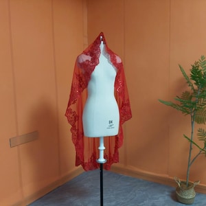 Può includere: Un velo di pizzo rosso è drappeggiato su un manichino bianco. Il velo ha un bordo smerlato e intricati motivi floreali. Il manichino è su un supporto nero, contro un muro arancione. Una pianta verde in un cesto intrecciato è a destra.