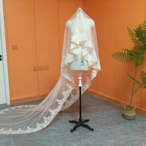 May include: A bridal veil on a mannequin. The veil is a soft ivory color with delicate lace trim along the edges and a long train. The mannequin is on a black stand, set against an orange wall with a small plant in a woven basket.