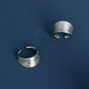 May include: Two silver-toned, open-ended rings with a textured, brushed metal finish. The rings have a wide band design and sit against a dark blue background. These adjustable rings are a simple, modern jewelry accessory.