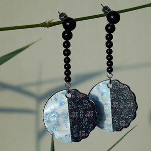May include: A pair of dangle earrings with black bead chains and circular pendants. Each pendant is split, with one half featuring a floral pattern in blue and white, and the other half a dark floral pattern with red accents.
