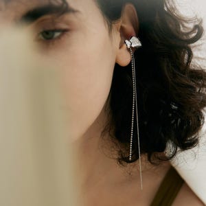 May include: A silver earring with a geometric design and a long, dangling chain of small beads. The earring is worn on the ear of a person with dark, curly hair. The earring is a modern and elegant piece of jewelry.
