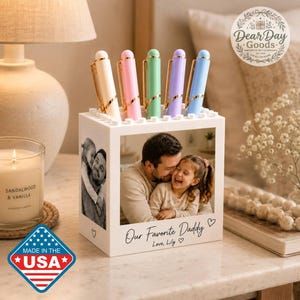 May include: A white square pen holder featuring a black and white photograph of a man and a child on the front, with the printed text Our Favorite Daddy Love, Lily. Five pastel-colored pens are inserted into the top of the blocky container.