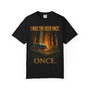 Peut inclure: T-shirt noir avec une scène de forêt représentant un loup et un cerf. Le texte "I WAS THE DEER ONCE. ONCE." est imprimé en or. Le motif est centré sur le devant du t-shirt.