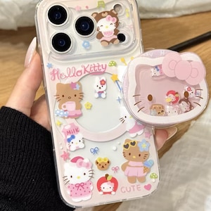 Puede incluir: Funda transparente para teléfono con Hello Kitty y otros personajes de dibujos animados. La funda tiene un diseño rosa y blanco con el texto "Hello Kitty" y "Cute". También se ve un agarre de teléfono emergente a juego.