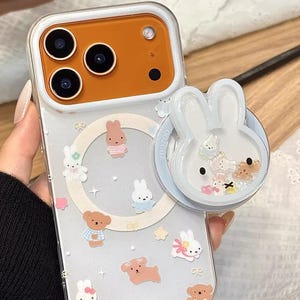 Puede incluir: Funda de teléfono transparente con borde blanco y zona de cámara naranja. La funda está decorada con conejos, osos y estrellas de dibujos animados. Se adjunta un agarre para teléfono blanco con forma de conejo. El texto "Cute V Case" está en la parte inferior.