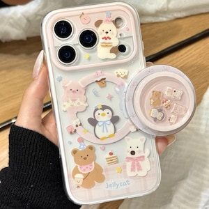 May include: A clear phone case with a pink border and a pop-up grip. The case features cartoon animal designs, including a pig, penguin, bear, and dog. The text "Jellycat" is visible on the case. The pop-up grip has a pink background with small bear designs.