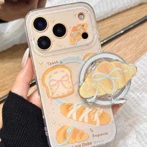 May include: Clear phone case with a bread-themed design. The case features illustrations of toast, a baguette, and a croissant, with the text "Toast", "Baguette", and "Lace Bake". A pop-up grip with a croissant design is attached.