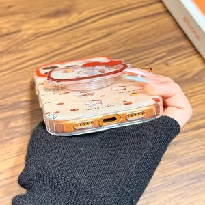 May include: A clear phone case with a Hello Kitty design, featuring a pop-up grip. The case has a gold-coloured border and is held by a person wearing a black jumper. The text "Cute V Case" is visible.