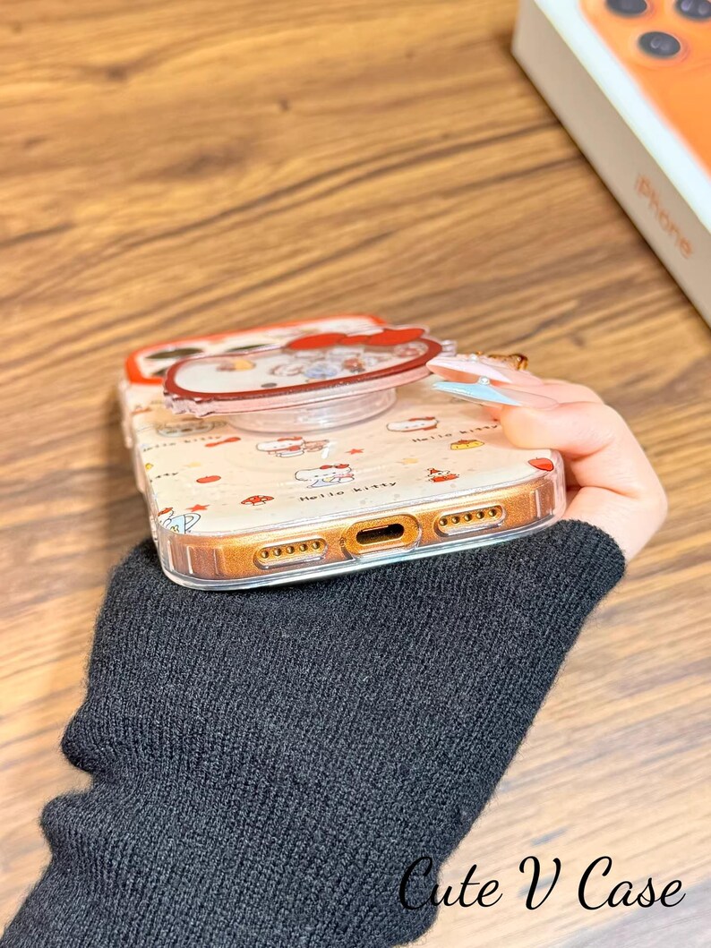 May include: A clear phone case with a Hello Kitty design, featuring a pop-up grip. The case has a gold-coloured border and is held by a person wearing a black jumper. The text "Cute V Case" is visible.