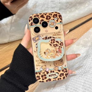 May include: A clear phone case with a leopard print design and a glitter-filled, heart-shaped pop socket. The case features cartoon illustrations of bears and Hello Kitty, with text that reads "Kitty." The case is held by a person.