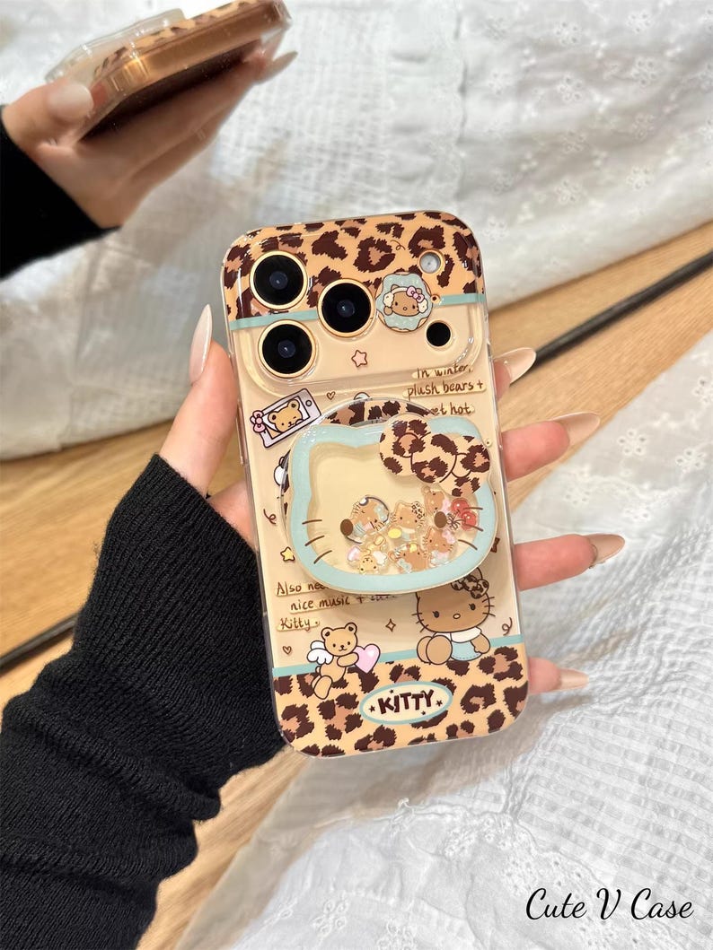 May include: A clear phone case with a leopard print design and a glitter-filled, heart-shaped pop socket. The case features cartoon illustrations of bears and Hello Kitty, with text that reads "Kitty." The case is held by a person.