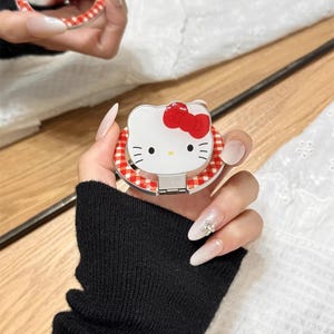 May include: A white compact mirror featuring a Hello Kitty design with a red bow and a red and white gingham border. The compact is held open, revealing the mirror. The text "Cute V Case" is visible.