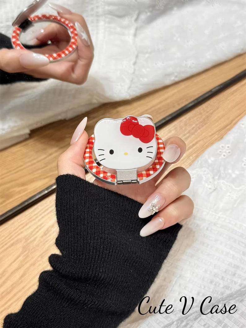 May include: A white compact mirror featuring a Hello Kitty design with a red bow and a red and white gingham border. The compact is held open, revealing the mirror. The text "Cute V Case" is visible.