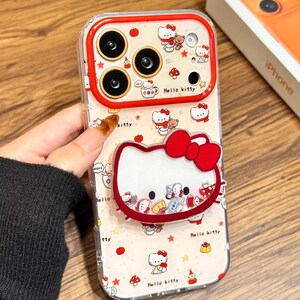 May include: Clear phone case with a red trim, decorated with a Hello Kitty design. The case features a repeating pattern of Hello Kitty, apples, and mushrooms. The text "Cute V Case" is visible.