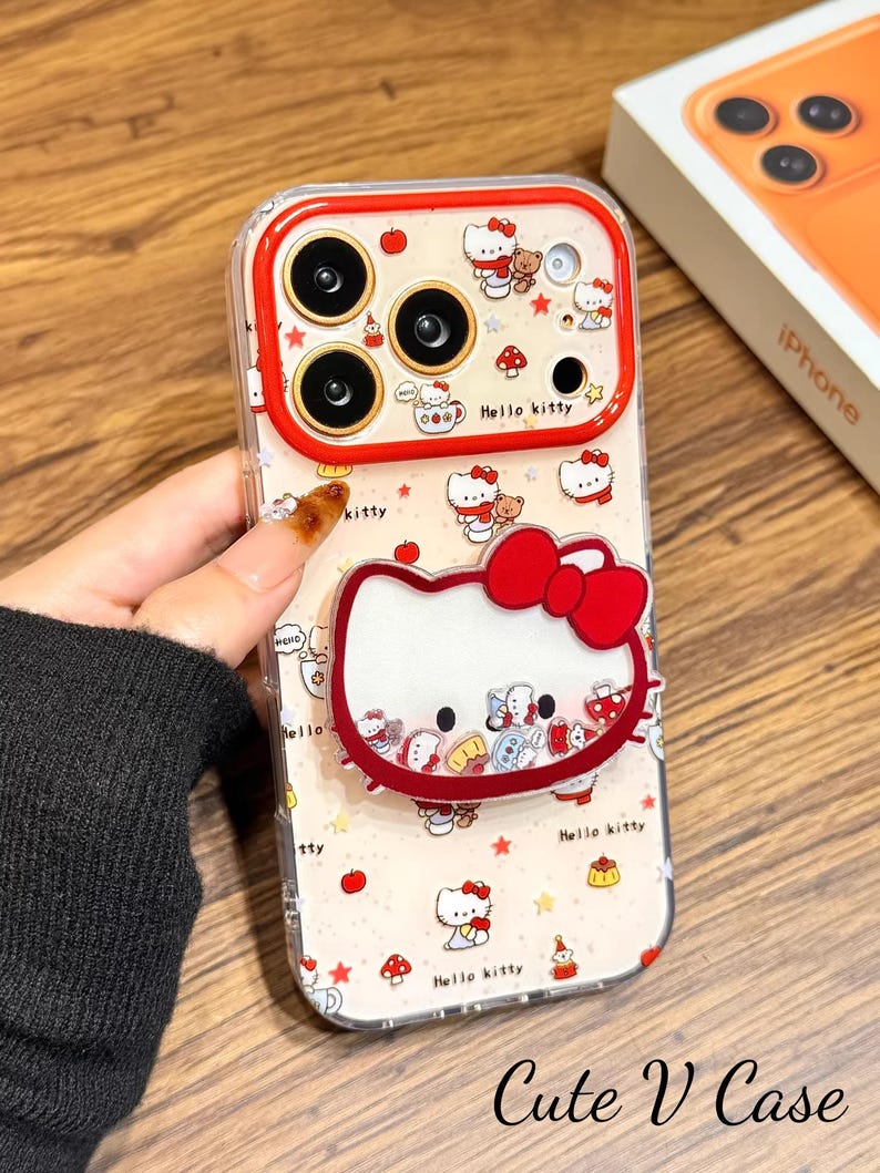 May include: Clear phone case with a red trim, decorated with a Hello Kitty design. The case features a repeating pattern of Hello Kitty, apples, and mushrooms. The text "Cute V Case" is visible.