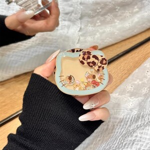 May include: A clear phone grip and a light blue phone case with a Hello Kitty design. The case has a leopard print bow and contains small, colorful figurines. The text "Cute V Case" is visible.