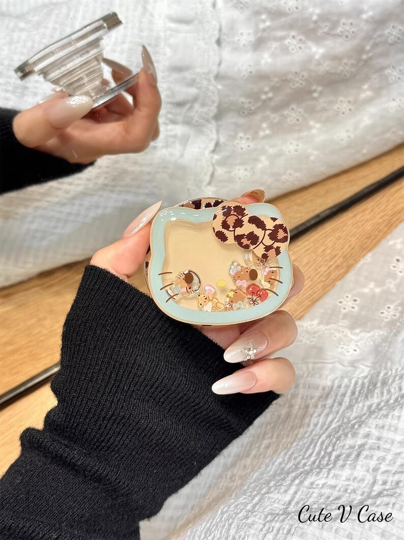 May include: A clear phone grip and a light blue phone case with a Hello Kitty design. The case has a leopard print bow and contains small, colorful figurines. The text "Cute V Case" is visible.