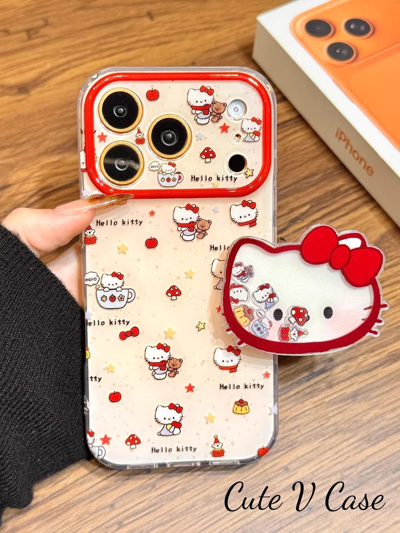 May include: A clear phone case with a red trim, decorated with a Hello Kitty pattern. The design includes Hello Kitty, teddy bears, mushrooms, and stars. A matching Hello Kitty phone grip is also shown.