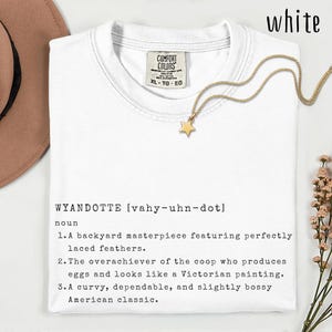 Pode incluir: Camiseta branca com a palavra "WYANDOTTE" e sua pronúncia impressas nela. A camisa também tem uma definição da palavra. Um colar de estrela dourada está drapeado na camisa.