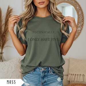 Op de afbeelding: Mosgroen t-shirt met de tekst "Technisch gezien, ik heb er maar vijf." Het shirt wordt gedragen met een blauwe spijkerbroek. Het model draagt het t-shirt.