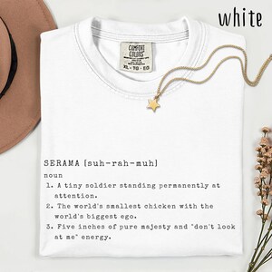 Puede incluir: Camiseta blanca con la palabra "SERAMA" y su pronunciación, junto con una definición. La camiseta también presenta un collar con colgante de estrella dorada. La camiseta está etiquetada XL-TG-EG.