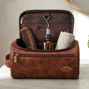 Könnte beinhalten: Eine braune Leder-Kulturtasche mit Reißverschluss, gefüllt mit einem Holzkamm, einer braunen Glasflasche und einem gerollten Tuch. Die Tasche hat eine goldene Berg-Grafik mit dem Namen "MARTIN" auf der Vorderseite.