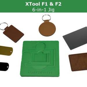 Può includere: Un assortimento di accessori per incisione laser, tra cui portachiavi, targhette e un jig verde con il testo "XTool F2 6-in-1 v1". I portachiavi sono marroni e circolari o rettangolari. Le targhette sono nere e dorate.