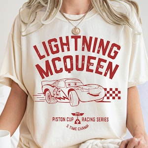 K&ouml;nnte beinhalten: Cremefarbenes T-Shirt mit roter Grafik von Lightning McQueen aus dem Film Cars. Das Design enth&auml;lt den Text "LIGHTNING MCQUEEN" &uuml;ber einer Illustration des Rennwagens, mit "Piston Cup Racing Series" und "5 Time Champ" darunter.