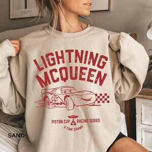K&ouml;nnte beinhalten: Sandfarbenes Sweatshirt mit einem roten, verblassten Aufdruck von Lightning McQueen, einem Rennwagen aus dem Film Cars. Das Design umfasst den Text "LIGHTNING MCQUEEN", eine Rennwagen-Illustration und "Piston Cup Racing Series S Time Champ".