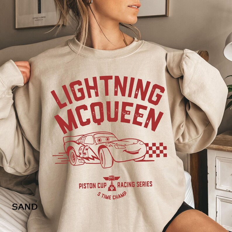 K&ouml;nnte beinhalten: Sandfarbenes Sweatshirt mit einem roten, verblassten Aufdruck von Lightning McQueen, einem Rennwagen aus dem Film Cars. Das Design umfasst den Text "LIGHTNING MCQUEEN", eine Rennwagen-Illustration und "Piston Cup Racing Series S Time Champ".