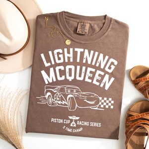 K&ouml;nnte beinhalten: Ein braunes T-Shirt mit dem Text "LIGHTNING MCQUEEN" und einer Grafik eines Rennwagens. Das Shirt tr&auml;gt auch den Text "PISTON CUP RACING SERIES S TIME CHAMP". Eine goldene Halskette, ein Hut und Sandalen sind ebenfalls im Bild.
