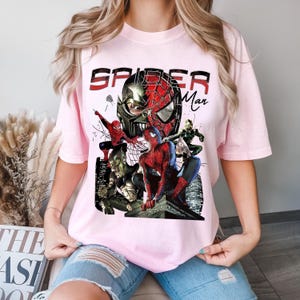 May include: Pale pink t-shirt with a Spider-Man graphic. The design features Spider-Man and other characters, with the word "SPIDER" in black and "Man" in red. The graphic has a comic book style, with various poses of Spider-Man and villains.