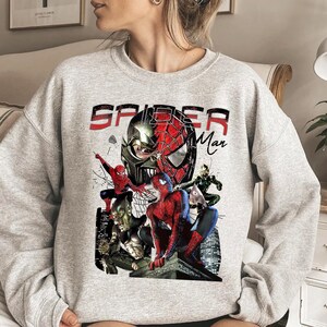 May include: A light grey sweatshirt with a Spider-Man graphic. The design features the words "Spider-Man" in red and black, and illustrations of Spider-Man and other characters from the Spider-Man universe.