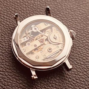 Könnte beinhalten: Nahaufnahme eines silberfarbenen Uhrengehäuses mit transparentem Zifferblatt, das die komplizierten Innenmechanismen zeigt. Die Uhr hat ein rundes Zifferblatt mit einer silbernen Lünette und einer strukturierten Krone. Die Uhr liegt auf einer dunkelbraunen Lederoberfläche.