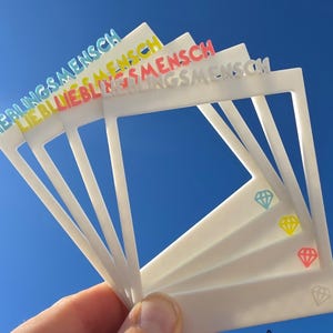 Könnte beinhalten: Ein Set aus weißen, rechteckigen Fotorahmen mit einer Aussparung in der Mitte. Jeder Rahmen zeigt oben das Wort LIEBLINGSMENSCH in erhabenen Buchstaben und unten rechts ein kleines Diamantsymbol in den Farben Blau, Gelb, Pink und Weiß.