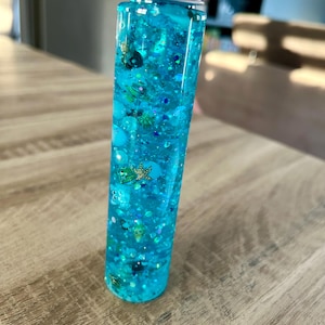 Könnte beinhalten: Eine transparente, zylindrische Sensorikflasche, gefüllt mit türkisfarbener Flüssigkeit, Glitzer und kleinen dekorativen Elementen. Die Flasche hat einen silbernen Deckel und enthält verschiedene Formen und Größen von blauen und grünen Verzierungen.