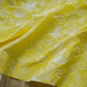 May include: Close-up of a yellow fabric with a floral pattern. The fabric has a textured surface with raised flower designs in a lighter yellow hue. The folds and shadows create depth.