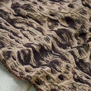 Könnte beinhalten: Nahaufnahme eines strukturierten Stoffes mit einem wirbelnden Muster in Braun- und Beigetönen. Der Stoff hat eine gekräuselte Textur, wobei sich das Licht von den erhabenen Bereichen reflektiert. Der Stoff ist über einem offenen Buch drapiert.