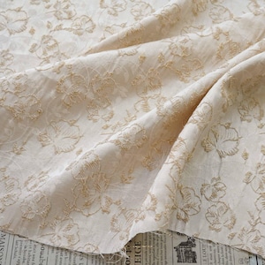 May include: Cream-colored fabric with a floral embroidered design. The fabric is draped, showing the texture and pattern of the embroidery. The floral pattern is in a gold color, creating a contrast against the cream background. The fabric appears to be soft and flowing.