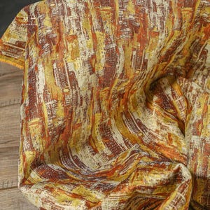 May include: A piece of fabric featuring an abstract pattern with vertical brushstroke-like textures in shades of golden yellow, burnt orange, and cream. The textile has a woven appearance with a visible grain and a small logo printed in the corner.