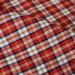 Può includere: Primo piano di un tessuto a quadri rosso, bianco e blu navy. Il tessuto presenta un classico motivo a scacchi con quadrati di dimensioni variabili. Il materiale è piegato, mostrando la trama e la profondità delle fibre tessute. I colori sono vivaci e il motivo è simmetrico.