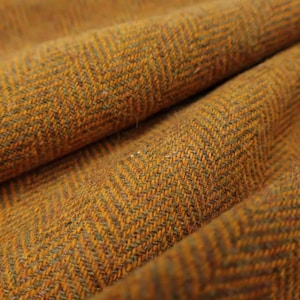 Puede incluir: Primer plano de una tela texturizada con un patrón de espiga. La tela es de un color marrón dorado cálido con sutiles acentos verde oscuro. El tejido crea una textura visual, con pliegues y sombras que añaden profundidad.
